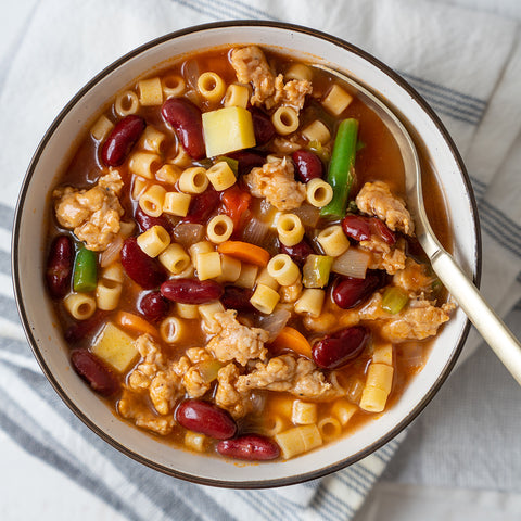 A bowl of minestrone soup with sausage, beans, and pasta, with a spoon on the side.