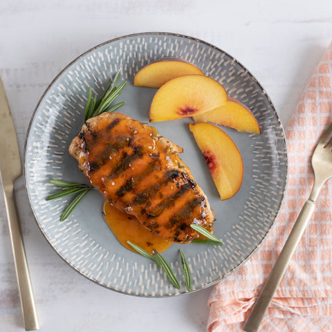 A plate with a grilled pork chop topped with peach sauce, alongside peach slices and a sprig of rosemary.